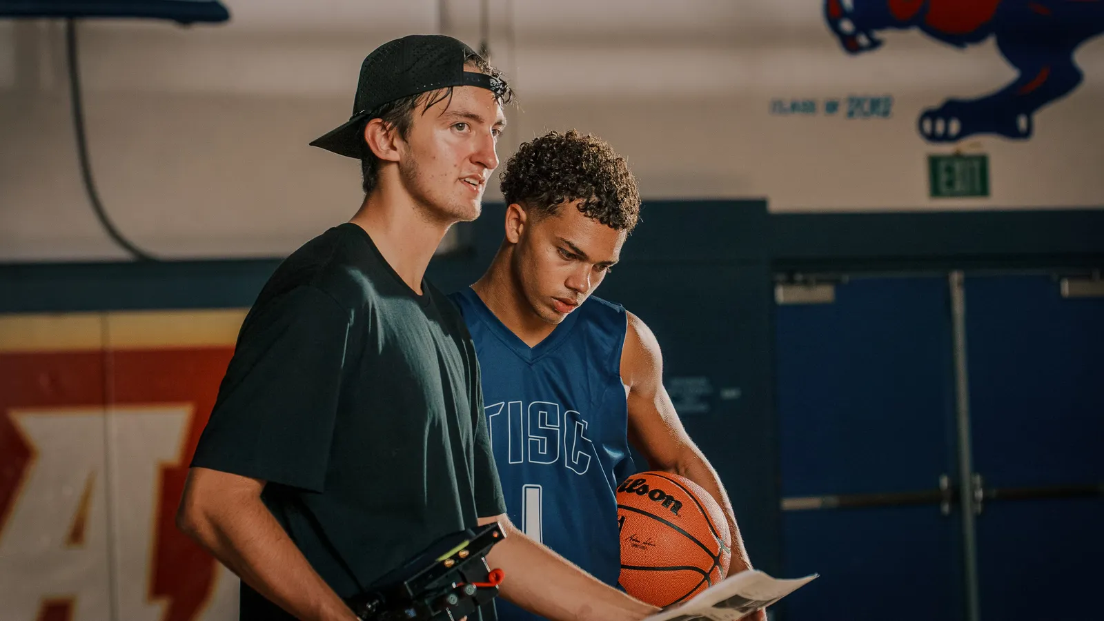 A director with a camera rig and a basketball player reviewing a shot list in a gym